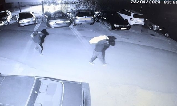 Czarno-biały kadr na tle zaparkowanych samochodów  widoczni sprawcy kradzieży,  którzy idą z workami na plecach